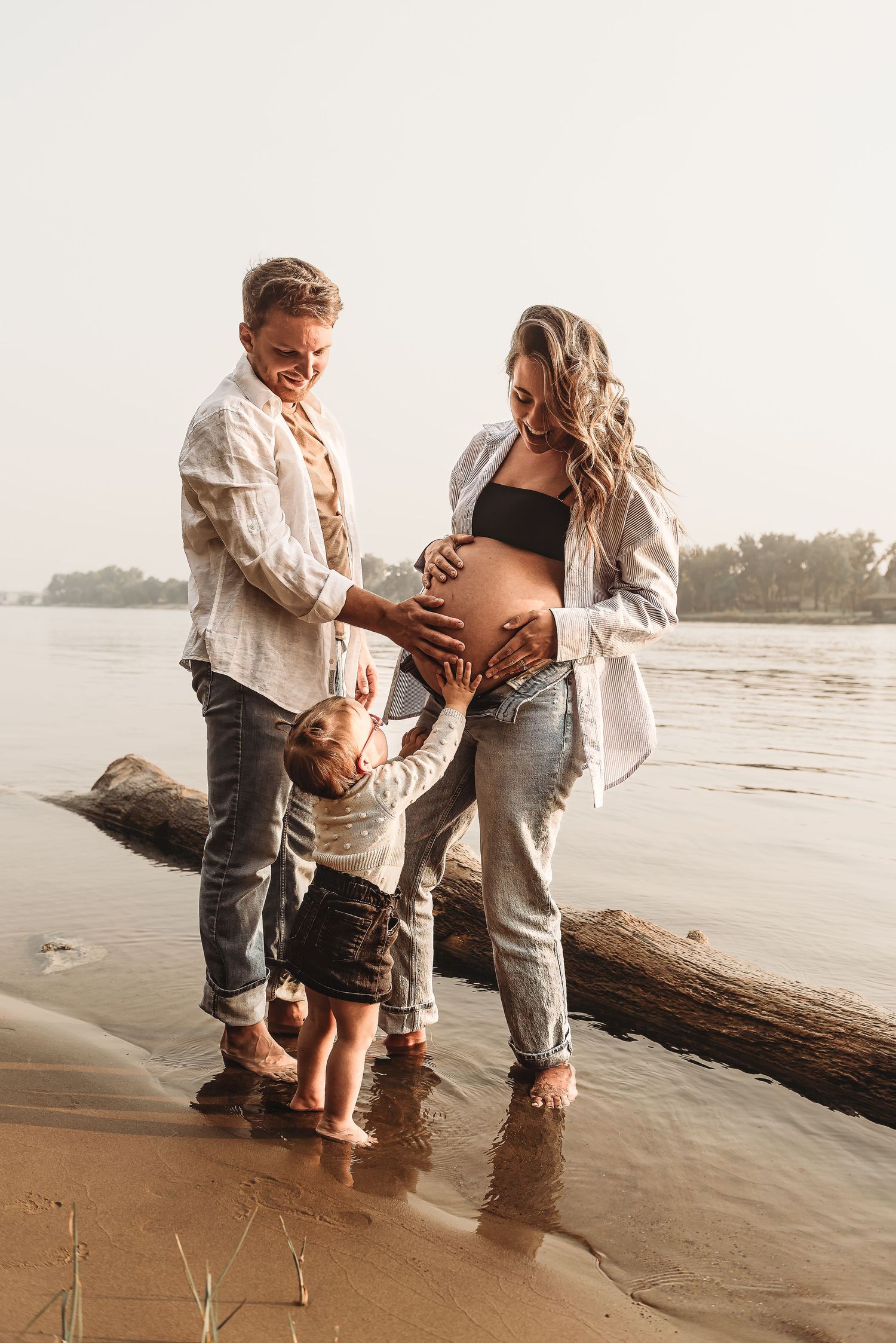 Expecting couple and toddler share intimate moment on beach at sunset.