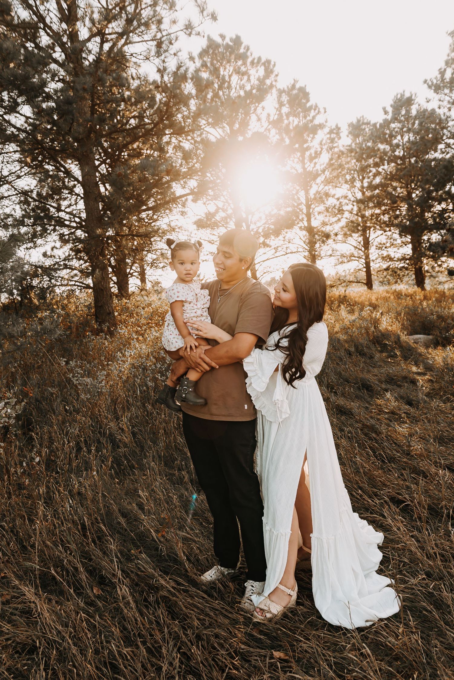Romantic couple poses in white dress and casual wear among pine trees at sunset.